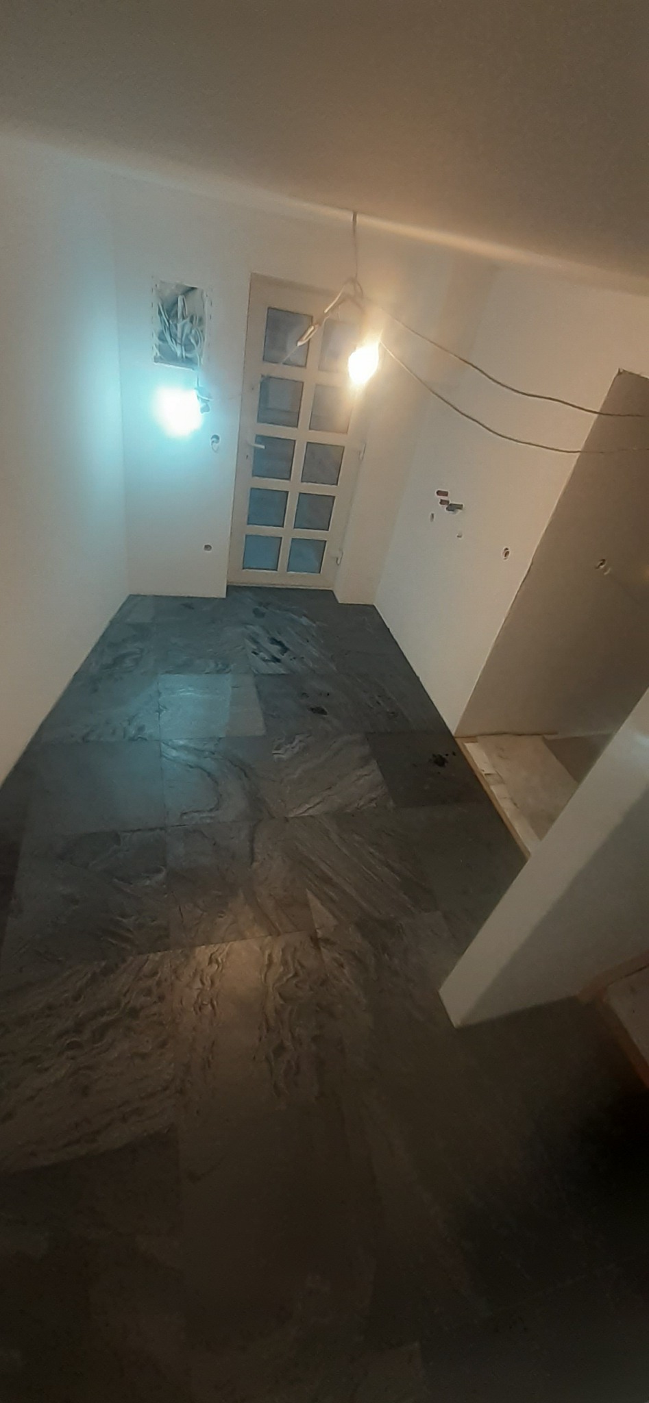 Ułożona ciemna, kamienna podłoga w pomieszczeniu z białymi ścianami, widoczne kable wystające ze ściany i niedokończone ościeżnice.