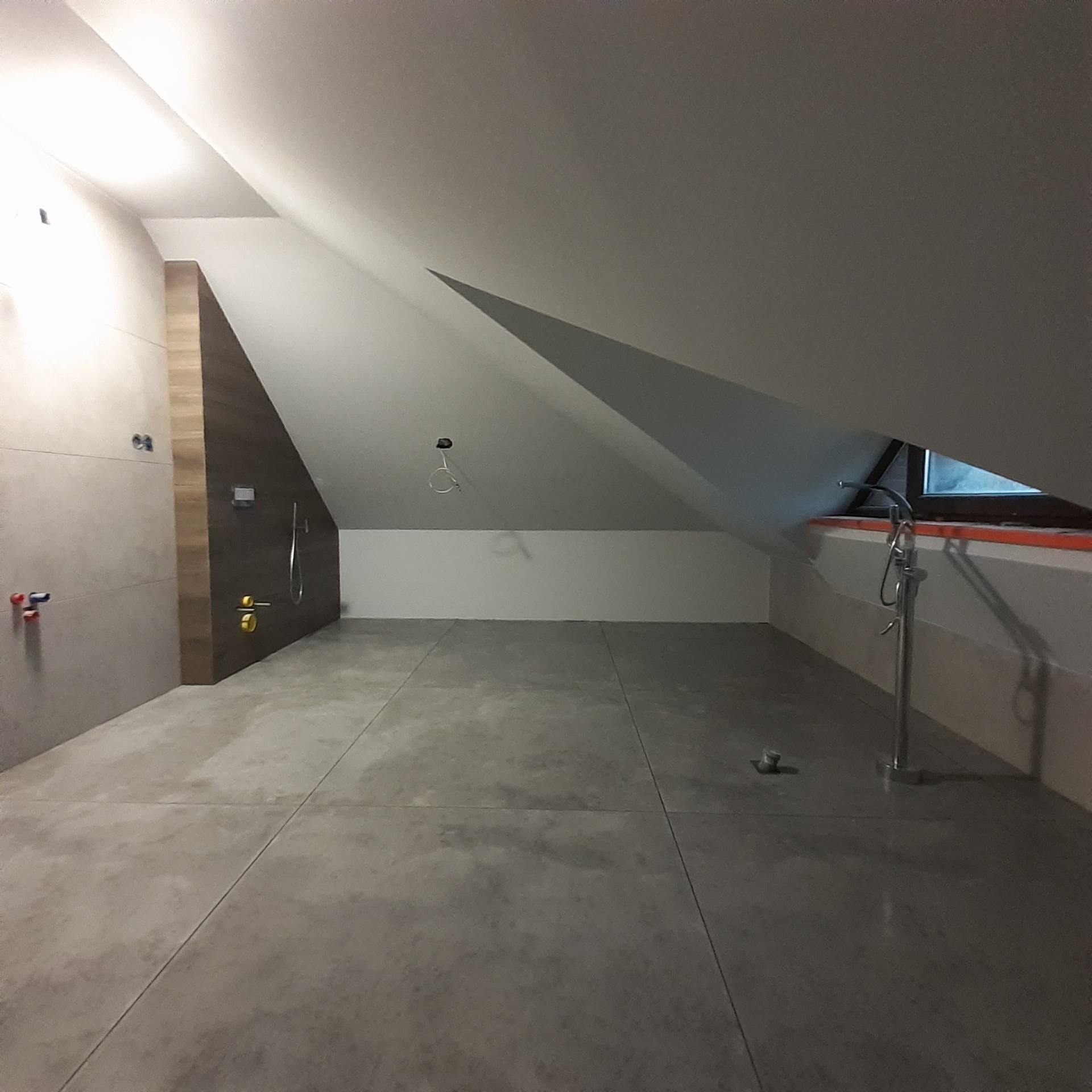 Surowa łazienka na poddaszu w trakcie wykańczania, widoczne płytki podłogowe, częściowo wykończona ściana prysznicowa z panelami imitującymi drewno, oraz instalacja wodna w pobliżu okna dachowego.