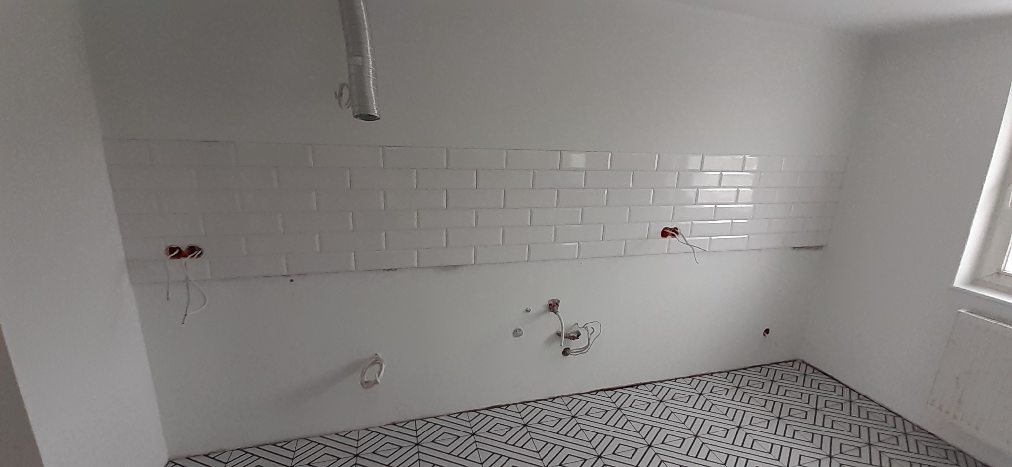 Białe, błyszczące płytki ułożone na ścianie, widoczne wystające kable elektryczne i rury, wzorzysta podłoga w czarno-białe geometryczne wzory, okno z grzejnikiem w rogu pomieszczenia.