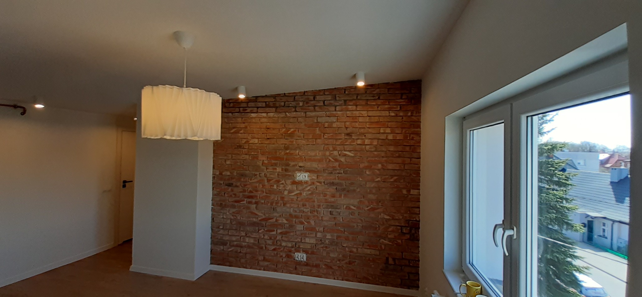 Pokój z fragmentem ściany wykończonej czerwoną cegłą, wiszącą lampą z plisowanym abażurem i oknem z widokiem na zewnątrz.