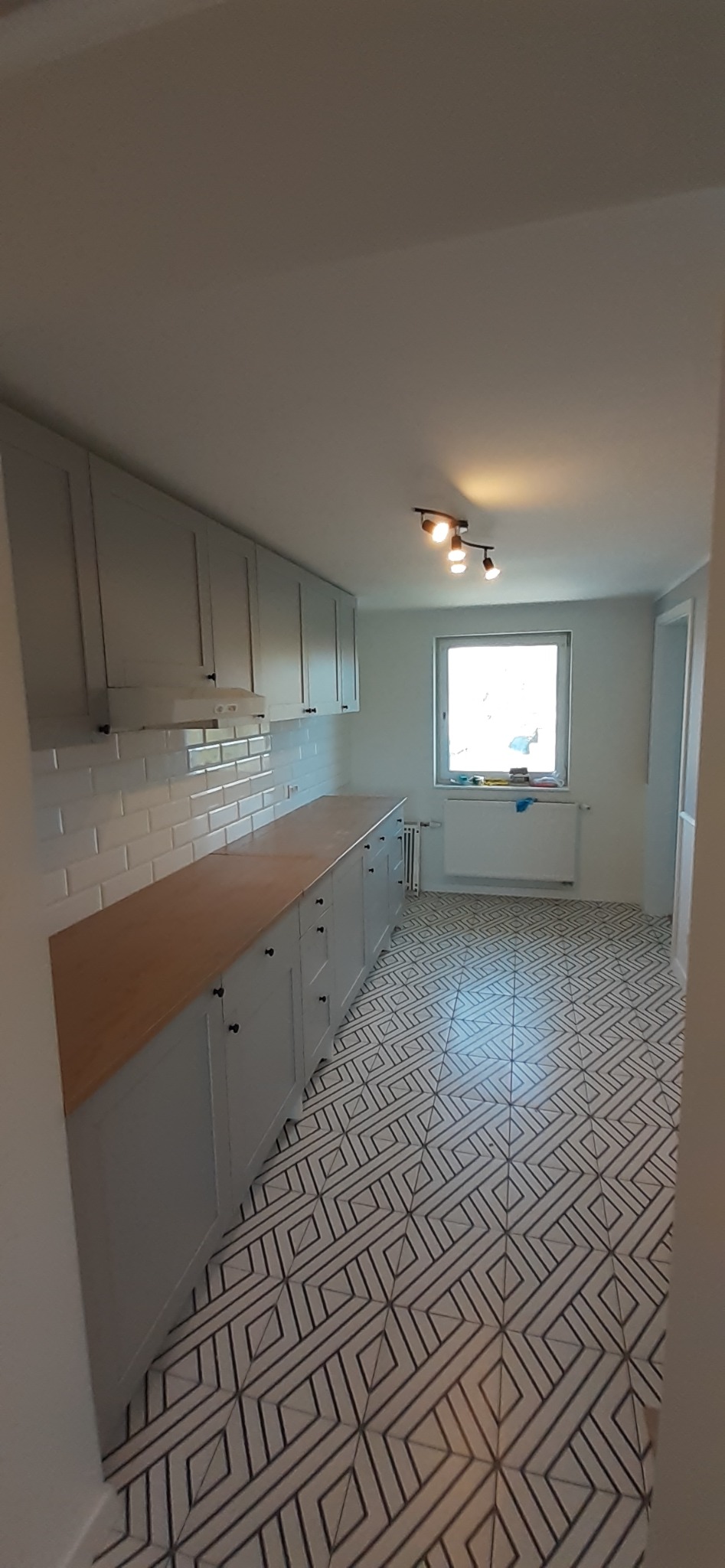 Jasna kuchnia po remoncie: szare szafki z drewnianym blatem, białe płytki na ścianie i wzorzysta podłoga z geometrycznym deseniem.