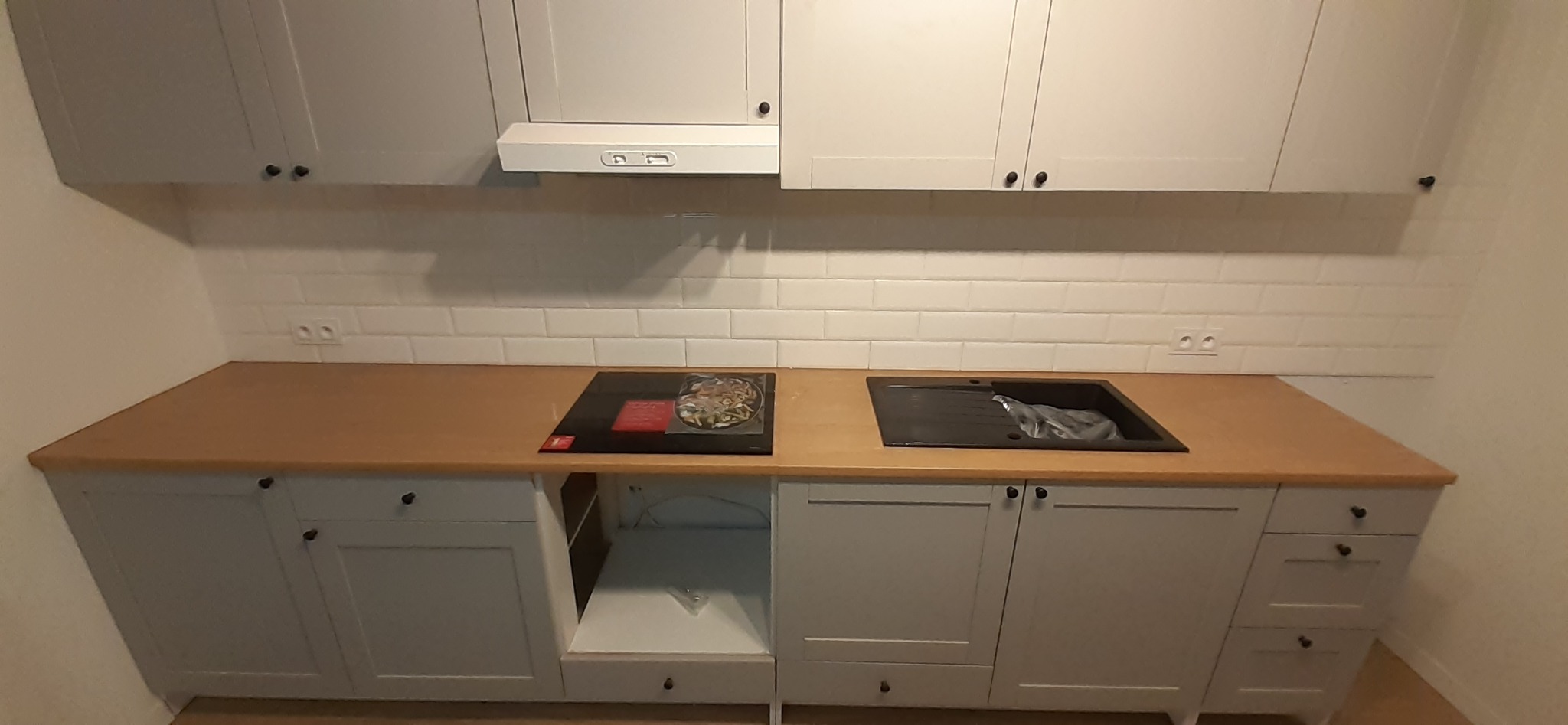 Jasnoszara zabudowa kuchenna z drewnianym blatem, białe kafelki na ścianie, czarny zlew i płyta indukcyjna z garnkiem.