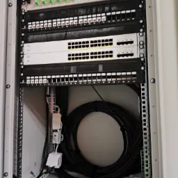 Szafa rack z przełącznikami sieciowymi Cisco, patch panelem i okablowaniem. Widoczne porty oznaczone numerami, kable uporządkowane wewnątrz szafy. Instalacja sieci komputerowej.