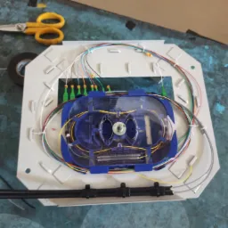 Otwarta skrzynka światłowodowa z widocznymi kablami, złączami i elementami montażowymi na tle stołu z niebieską, popękaną powierzchnią. Obok leżą nożyczki z żółtymi uchwytami i rolka czarnej taśmy...