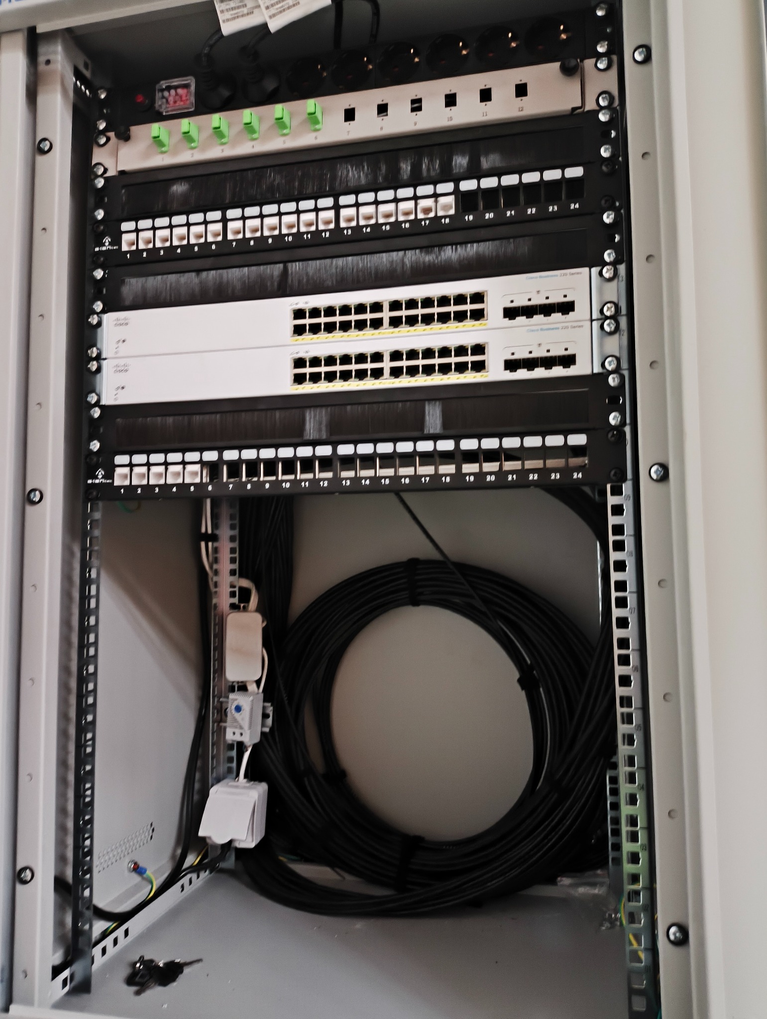 Szafa rack z przełącznikami sieciowymi Cisco, patch panelem i okablowaniem. Widoczne porty oznaczone numerami, kable uporządkowane wewnątrz szafy. Instalacja sieci komputerowej.