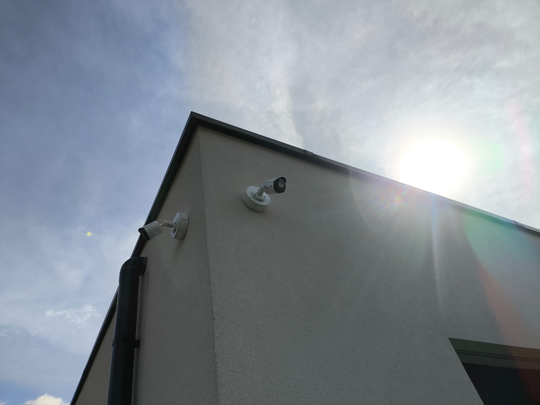 Dwie białe kamery monitoringu na rogu budynku z szarą elewacją, skierowane w różne strony, na tle jasnego, zachmurzonego nieba i słońca.