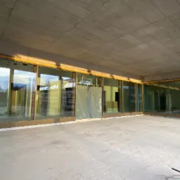 Wnętrze surowego budynku z panoramicznymi oknami w złotych ramach, jedno okno zasłonięte folią ochronną, betonowy sufit i podłoga w trakcie wykańczania.