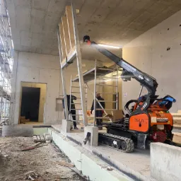 Montaż konstrukcji stalowej przy użyciu podnośnika gąsienicowego w surowym wnętrzu budynku. Widoczne rusztowania, betonowe ściany i podłoże w trakcie prac budowlanych.