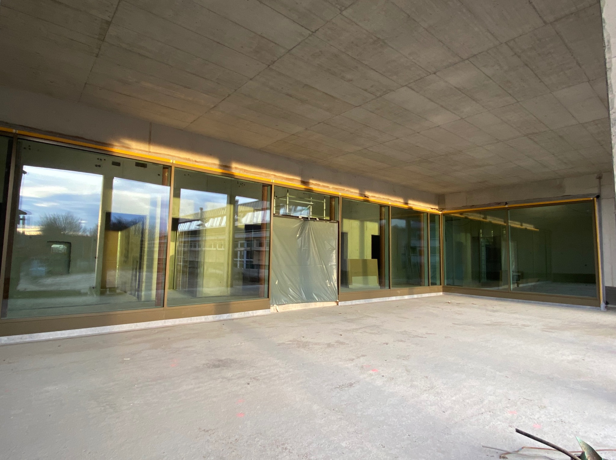 Wnętrze surowego budynku z panoramicznymi oknami w złotych ramach, jedno okno zasłonięte folią ochronną, betonowy sufit i podłoga w trakcie wykańczania.