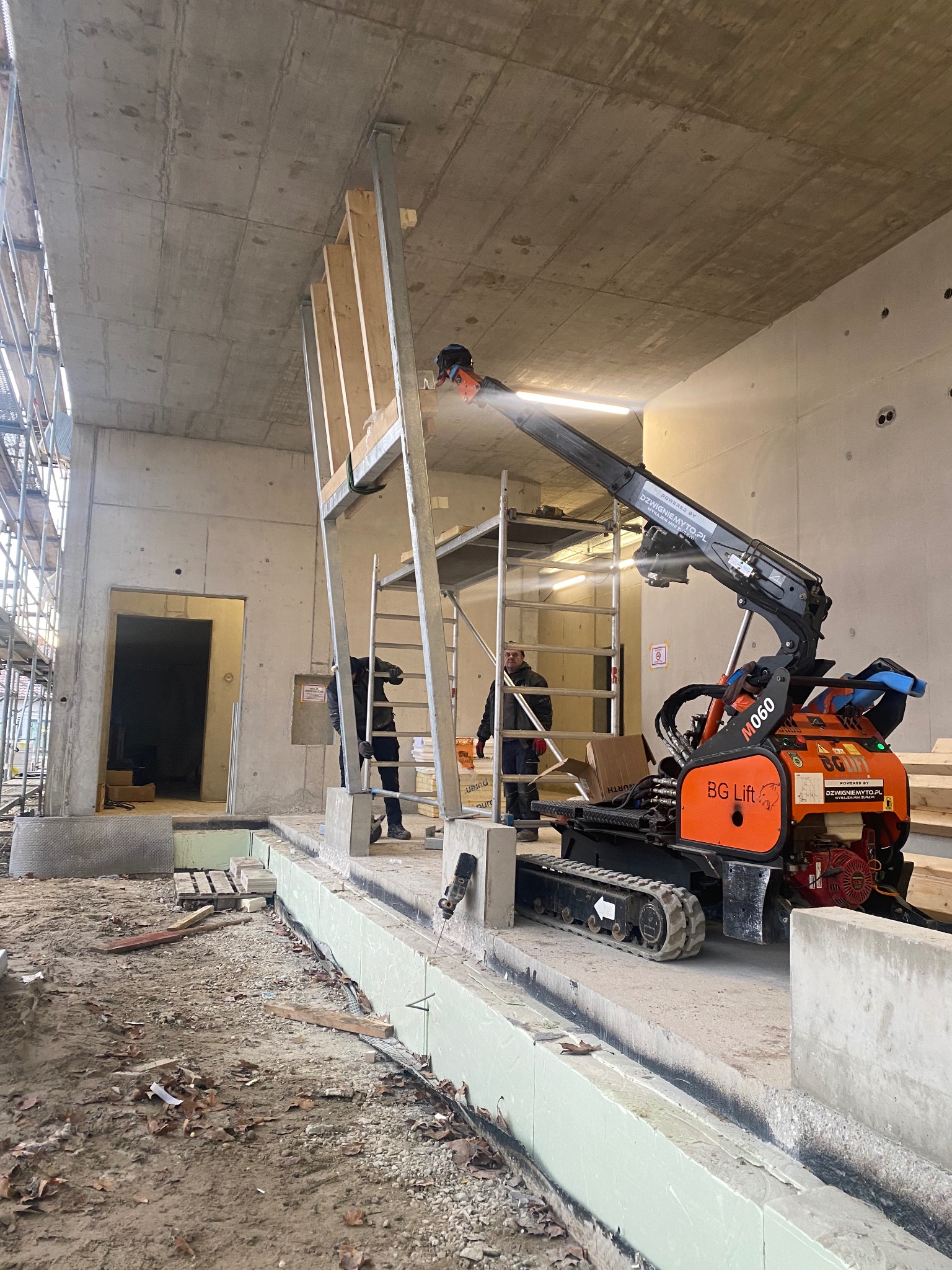 Montaż konstrukcji stalowej przy użyciu podnośnika gąsienicowego w surowym wnętrzu budynku. Widoczne rusztowania, betonowe ściany i podłoże w trakcie prac budowlanych.