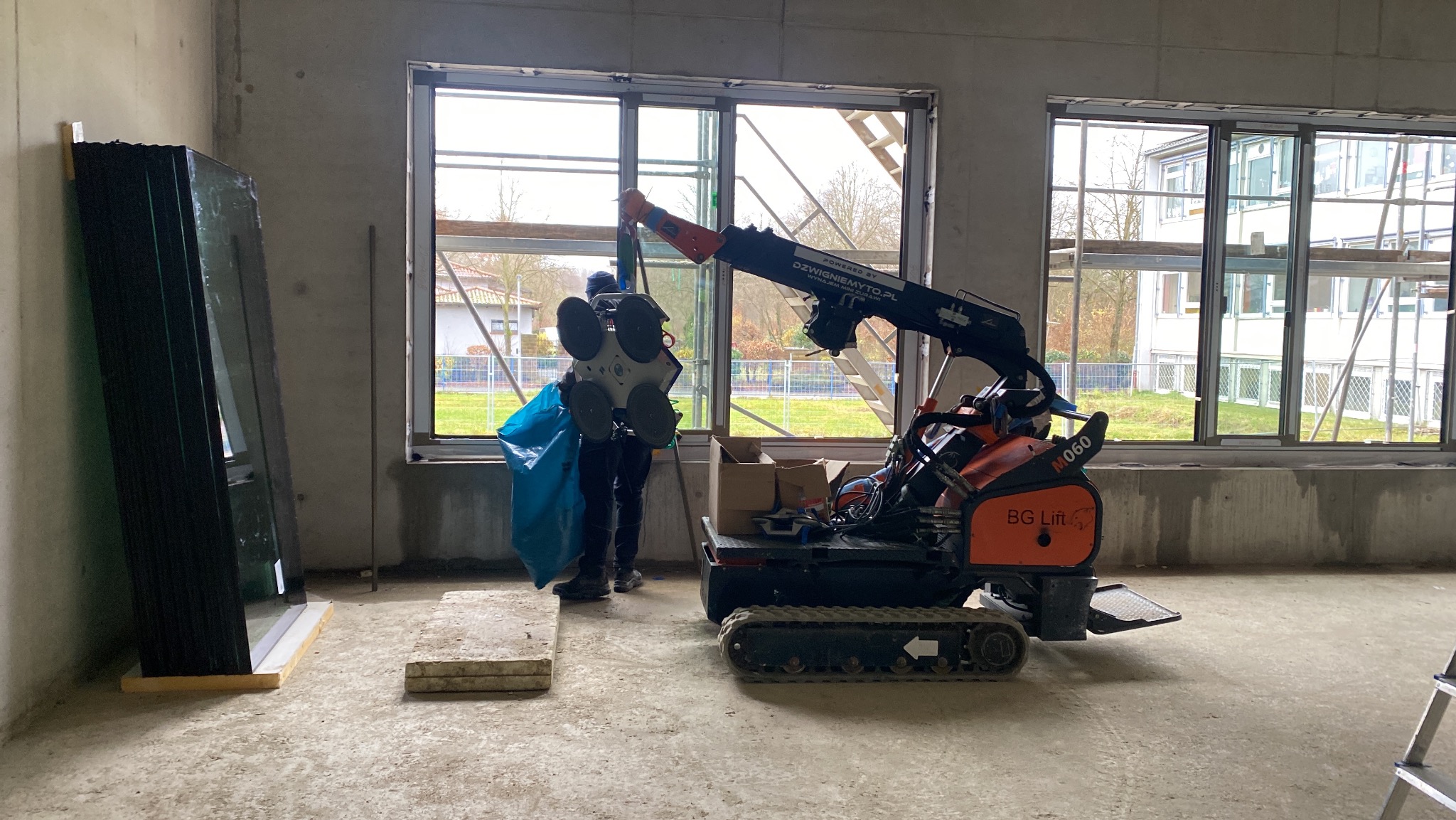 Instalacja okna z użyciem mini dźwigu BG Lift w surowym wnętrzu budynku; w tle widoczne okna i rusztowania.