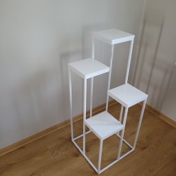 Kamil Jasztal - Trójstopniowy, biały, metalowy kwietnik w minimalistycznym stylu, ustawiony na parkiecie przy jasnej ścianie.