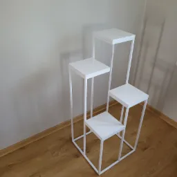 Trójstopniowy, biały, metalowy kwietnik w minimalistycznym stylu, ustawiony na parkiecie przy jasnej ścianie.