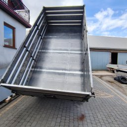 Kamil Jasztal - Podniesiona metalowa paka wywrotki z widocznymi spawami, umieszczona na kostce brukowej, w tle budynek z blachy falistej i zaparkowany żółty pojazd.