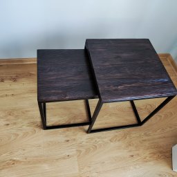 Kamil Jasztal - Dwa stoliki kawowe typu nesting, z ciemnobrązowymi blatami o widocznym usłojeniu i czarnymi, metalowymi nogami, ustawione na podłodze z paneli imitujących drewno, na tle białej ściany.