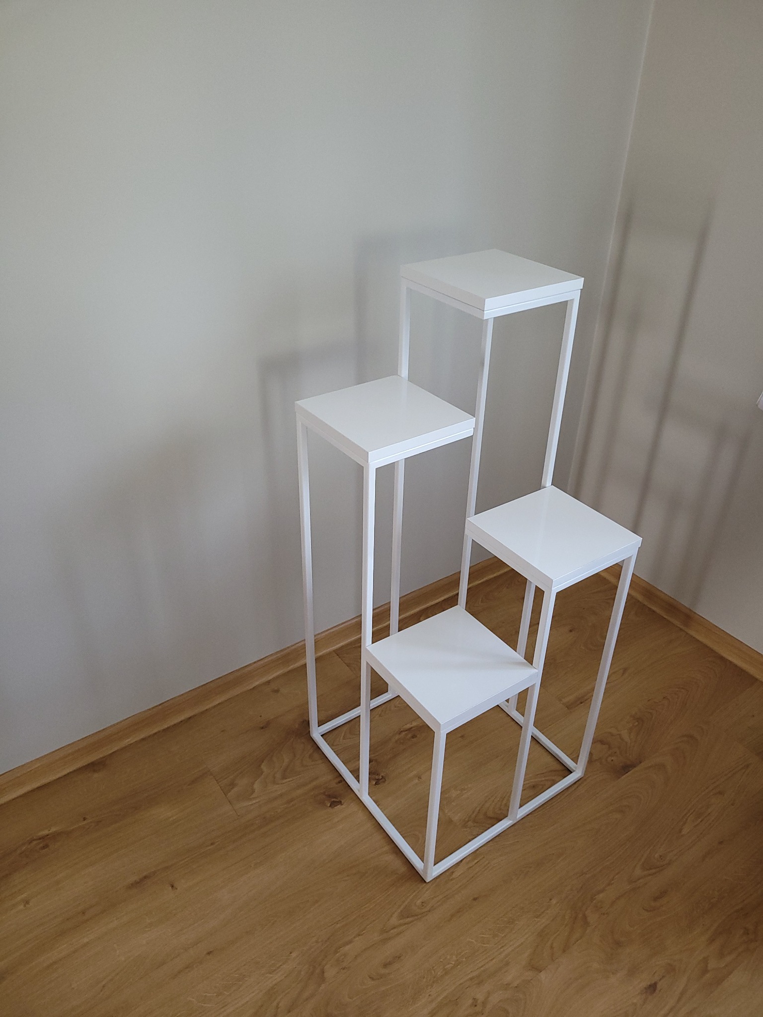 Trójstopniowy, biały, metalowy kwietnik w minimalistycznym stylu, ustawiony na parkiecie przy jasnej ścianie.