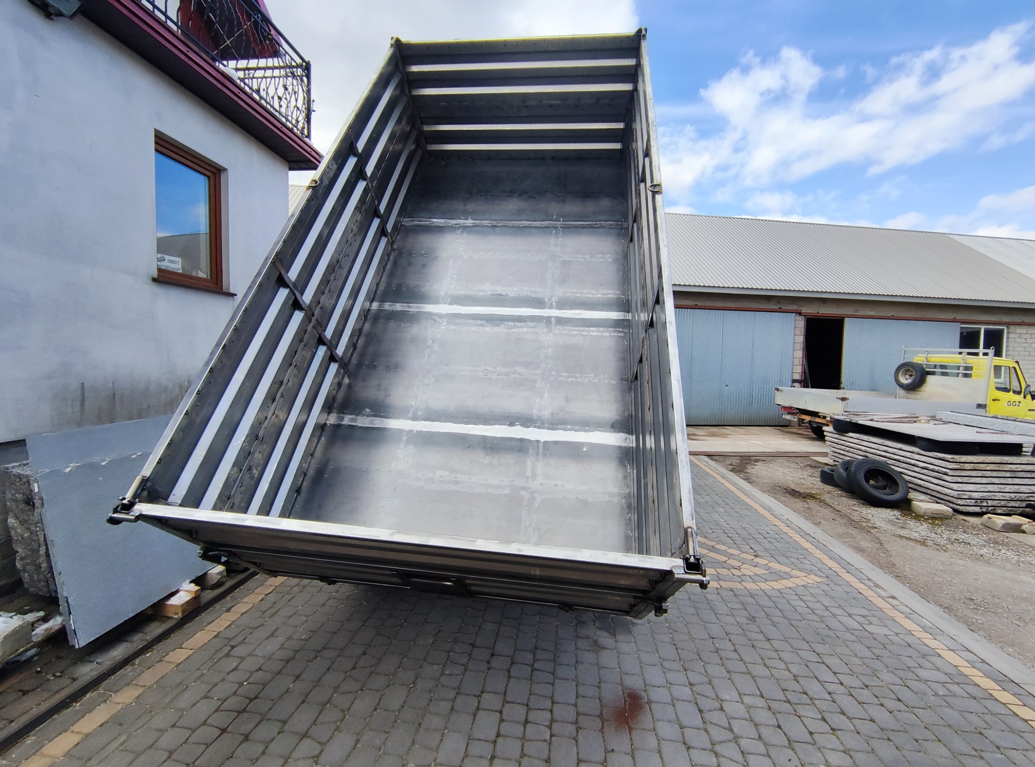 Podniesiona metalowa paka wywrotki z widocznymi spawami, umieszczona na kostce brukowej, w tle budynek z blachy falistej i zaparkowany żółty pojazd.