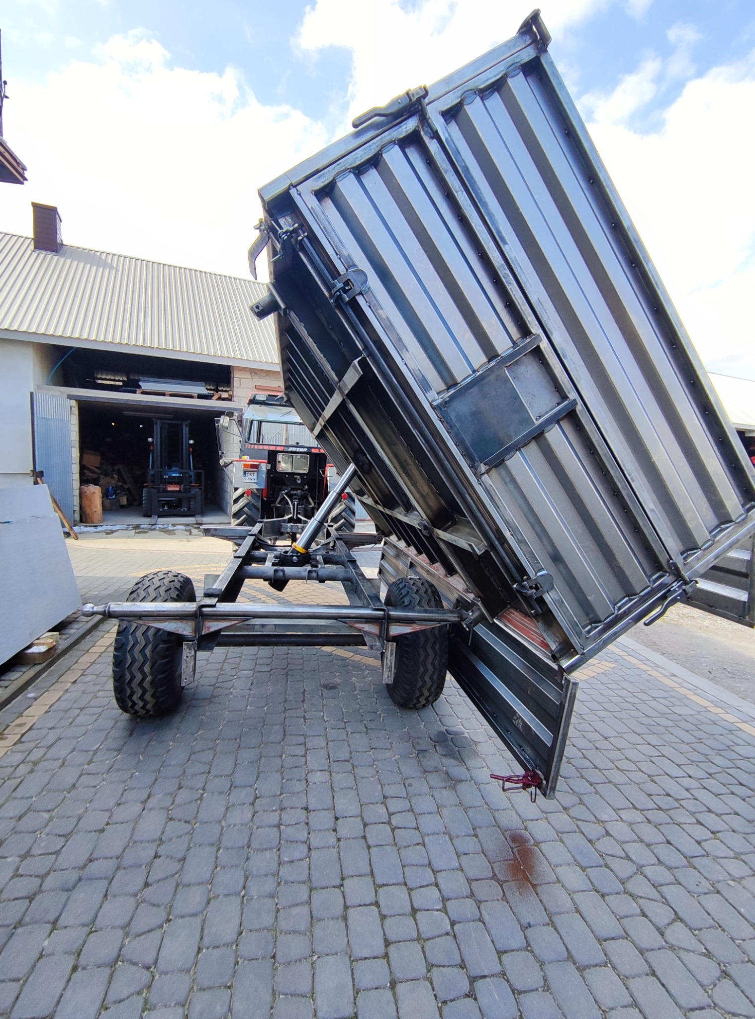 Metalowa przyczepa rolnicza z podniesioną skrzynią ładunkową, widoczny mechanizm podnoszenia, opony terenowe i ciągniki w tle.