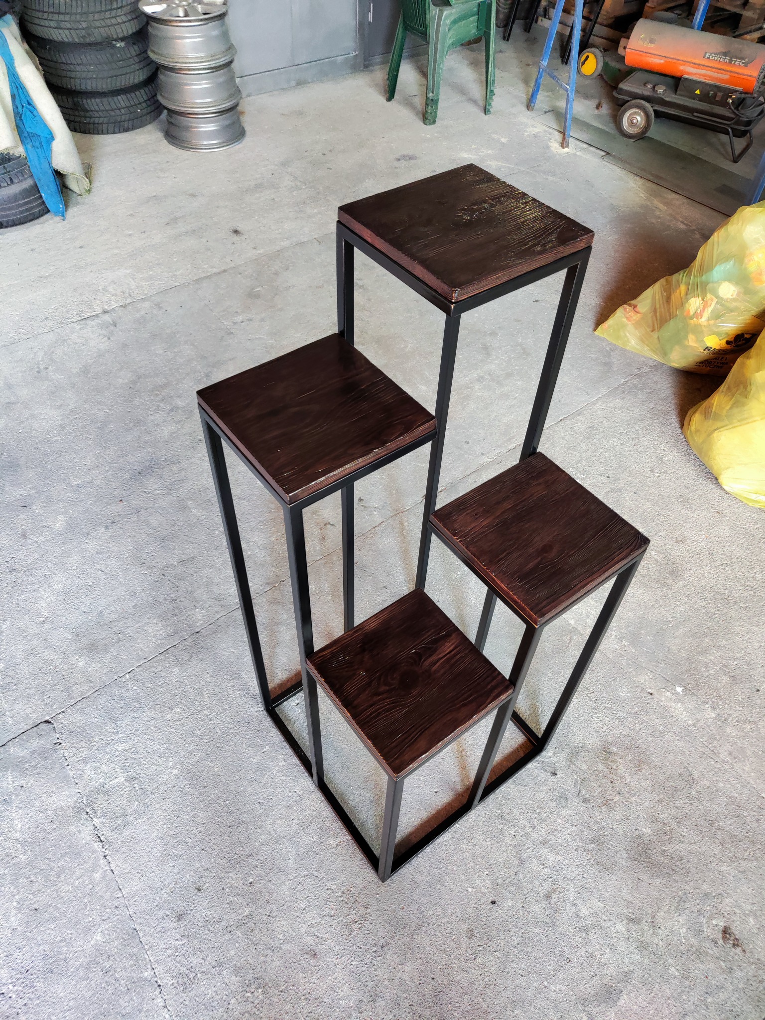 Cztery drewniane, lakierowane stołki o różnych wysokościach z czarnymi metalowymi ramami, ustawione schodkowo na betonowej podłodze warsztatu, w tle opony, butla gazowa i worki na śmieci.