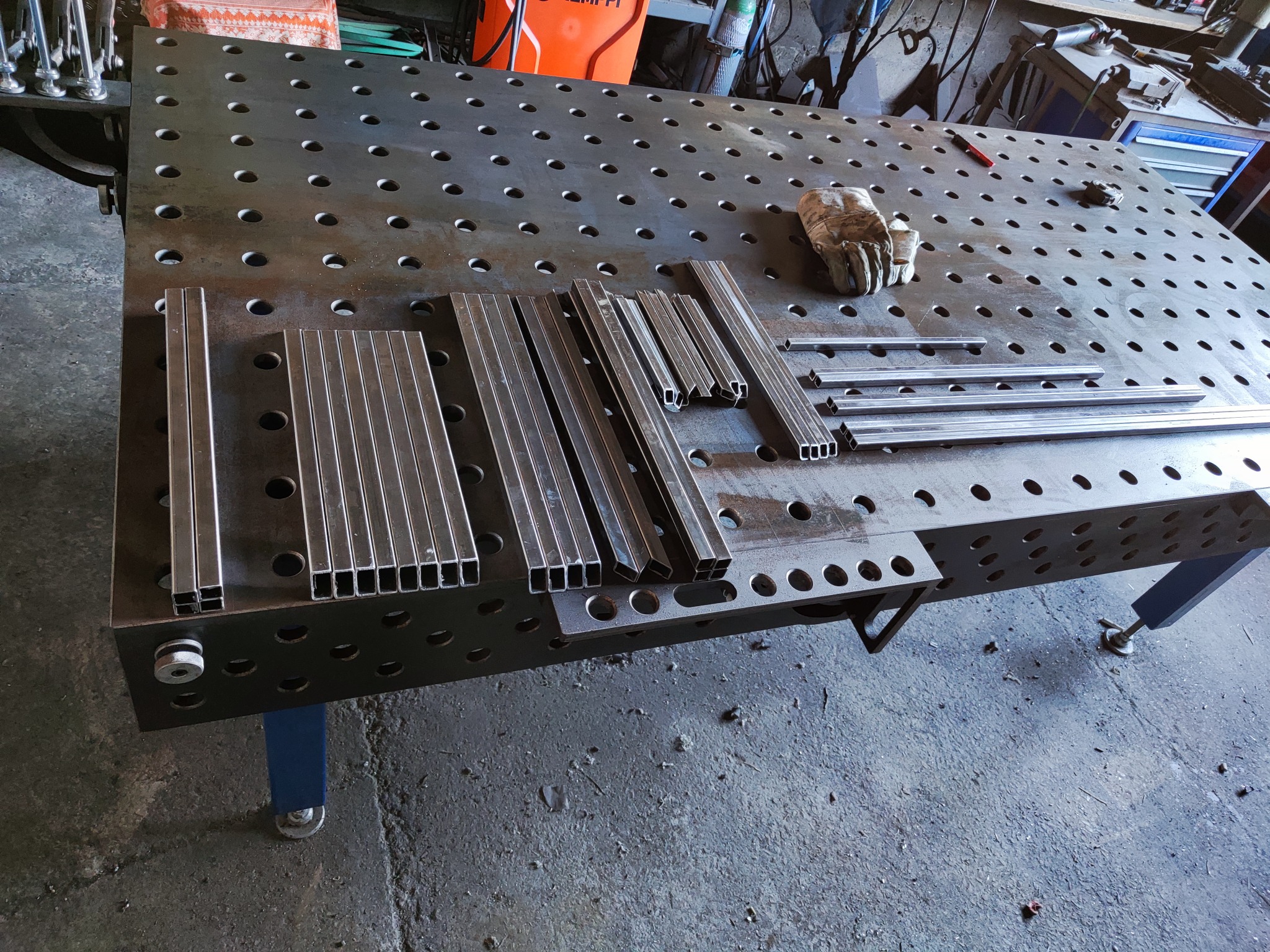 Przygotowane profile stalowe o przekroju kwadratowym i prostokątnym, ułożone na perforowanym stole spawalniczym, obok leży rękawica robocza