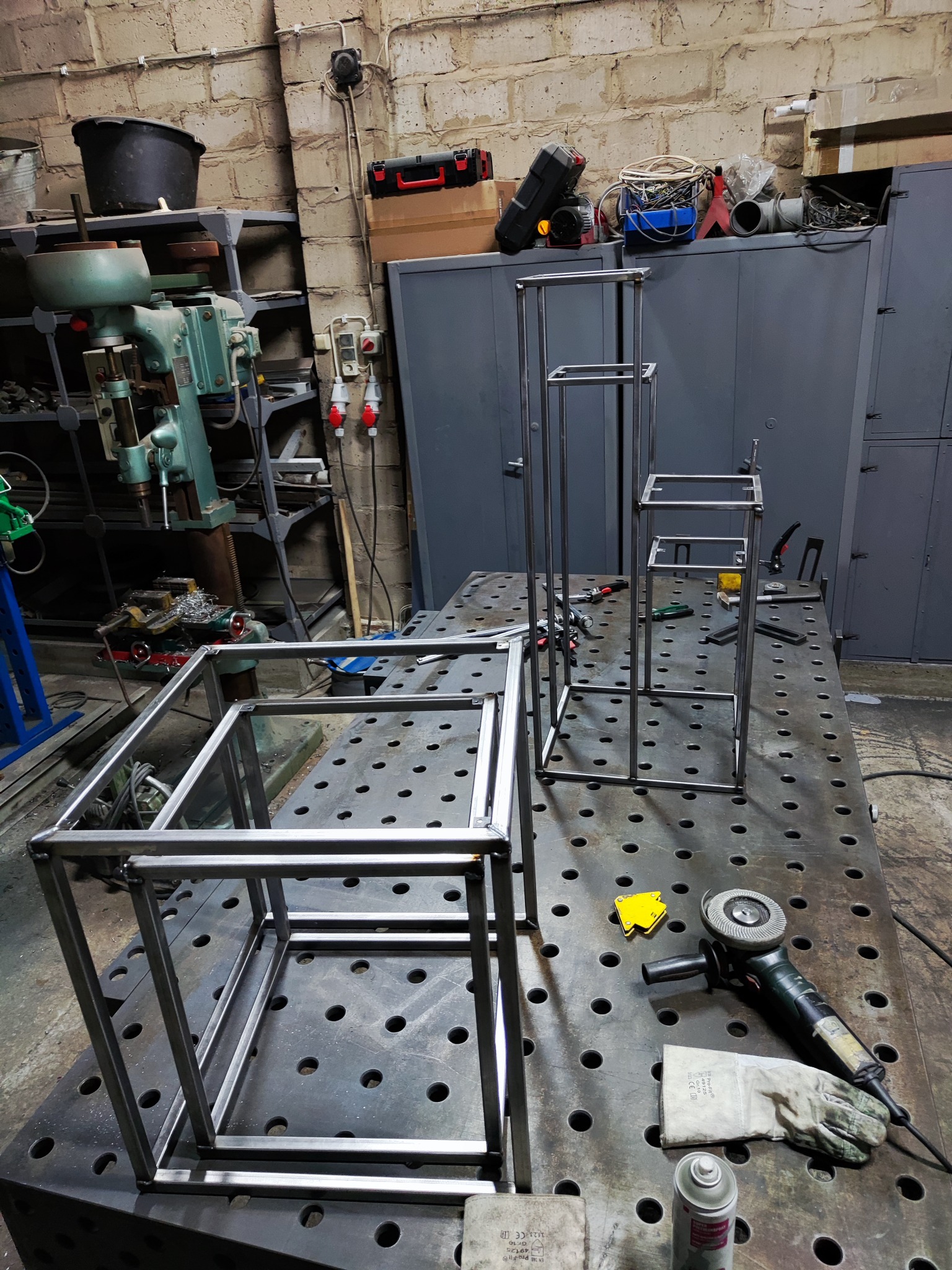 Metalowe konstrukcje szkieletowe, prawdopodobnie kwietniki, w różnej fazie wykonania, leżą na perforowanym stole warsztatowym w warsztacie, obok szlifierka kątowa i rękawica ochronna.