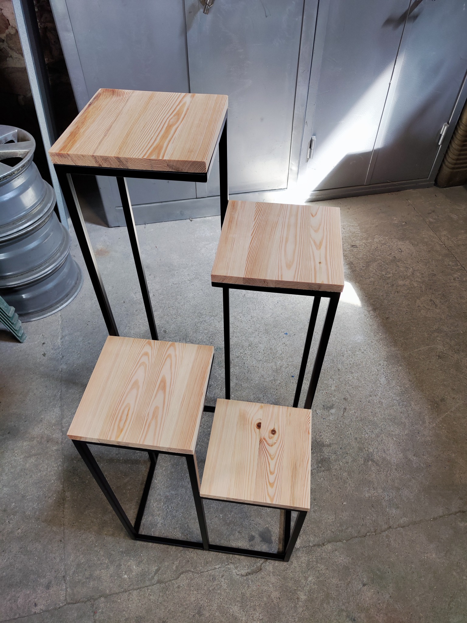 Czterostopniowy kwietnik z jasnego drewna o widocznym usłojeniu, z czarnymi, spawanymi, metalowymi nogami, na tle betonowej podłogi i metalowej szafy.