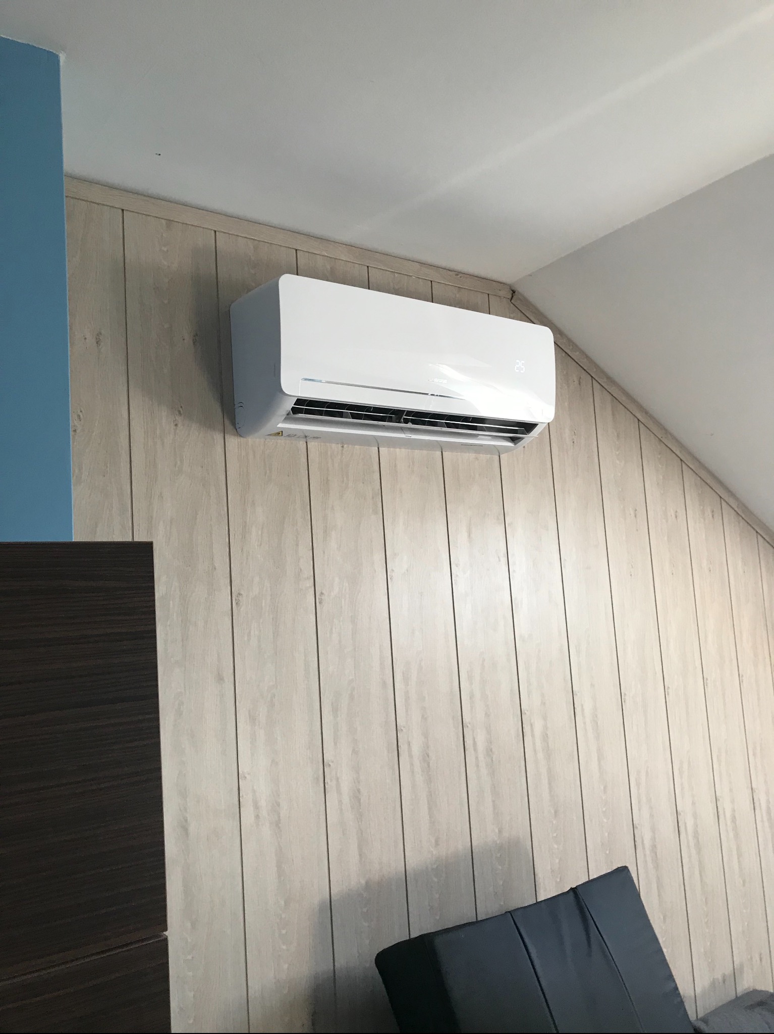 Biała jednostka wewnętrzna klimatyzatora zamontowana na ścianie z paneli imitujących drewno w pokoju z niebieskim akcentem kolorystycznym.