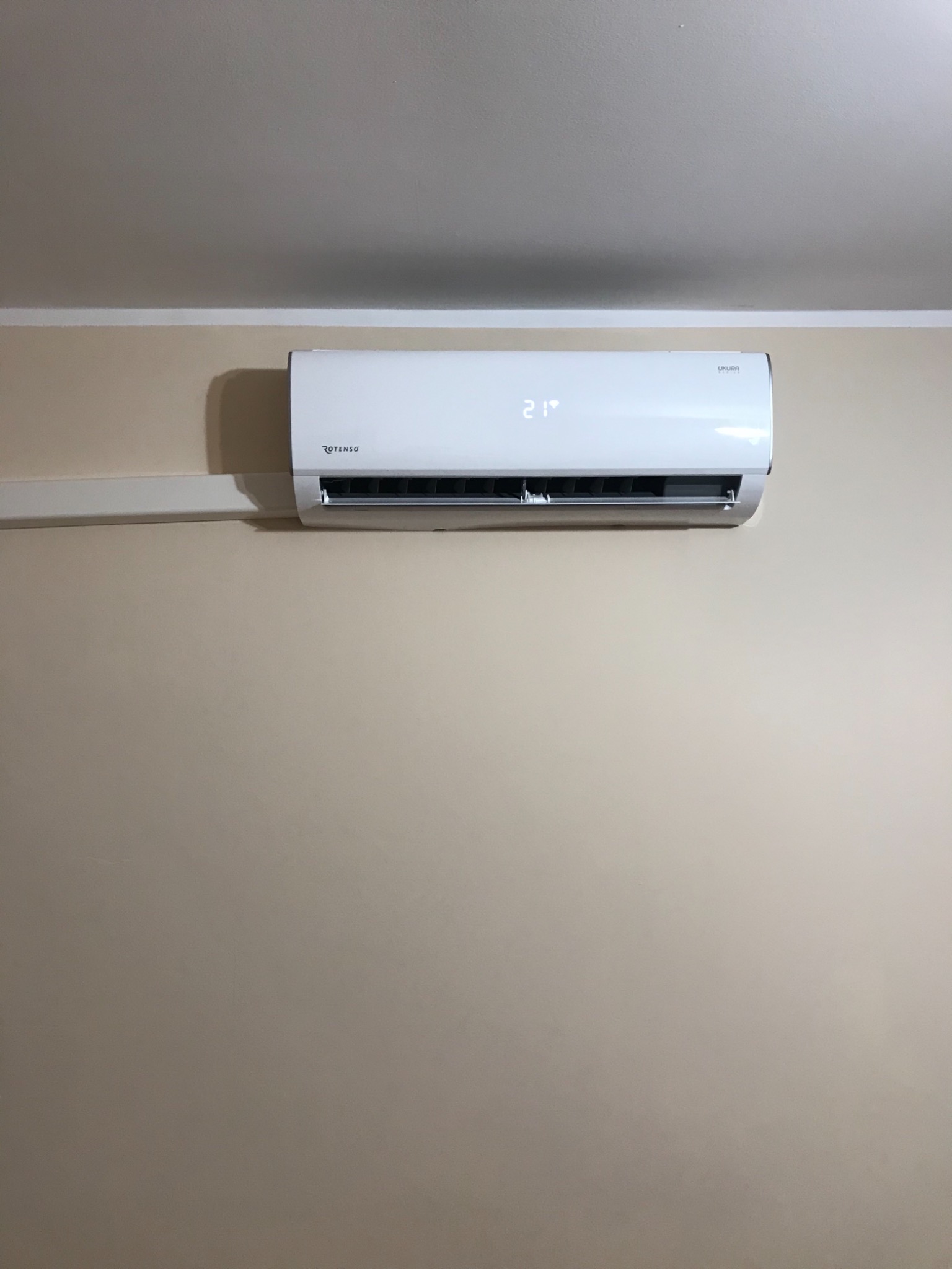Biały klimatyzator Rotenso Ukura zamontowany na beżowej ścianie, wyświetlający temperaturę 21 stopni Celsjusza.