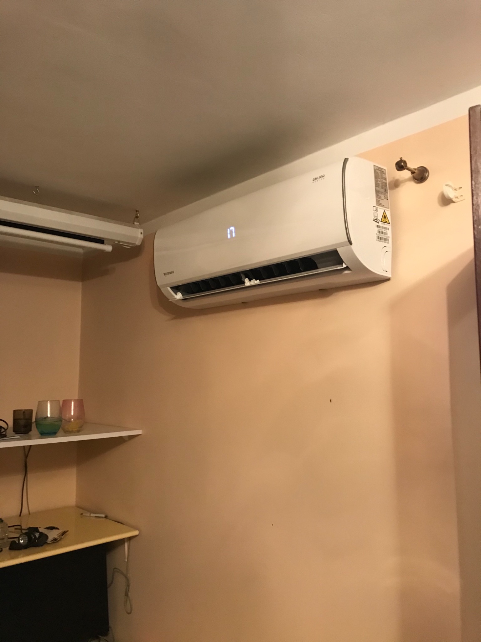 Biały klimatyzator Rotenso zamontowany na ścianie w pomieszczeniu o beżowych ścianach, wyświetlający temperaturę 17 stopni Celsjusza, obok widoczny drugi klimatyzator i półka z kolorowymi szklankami.