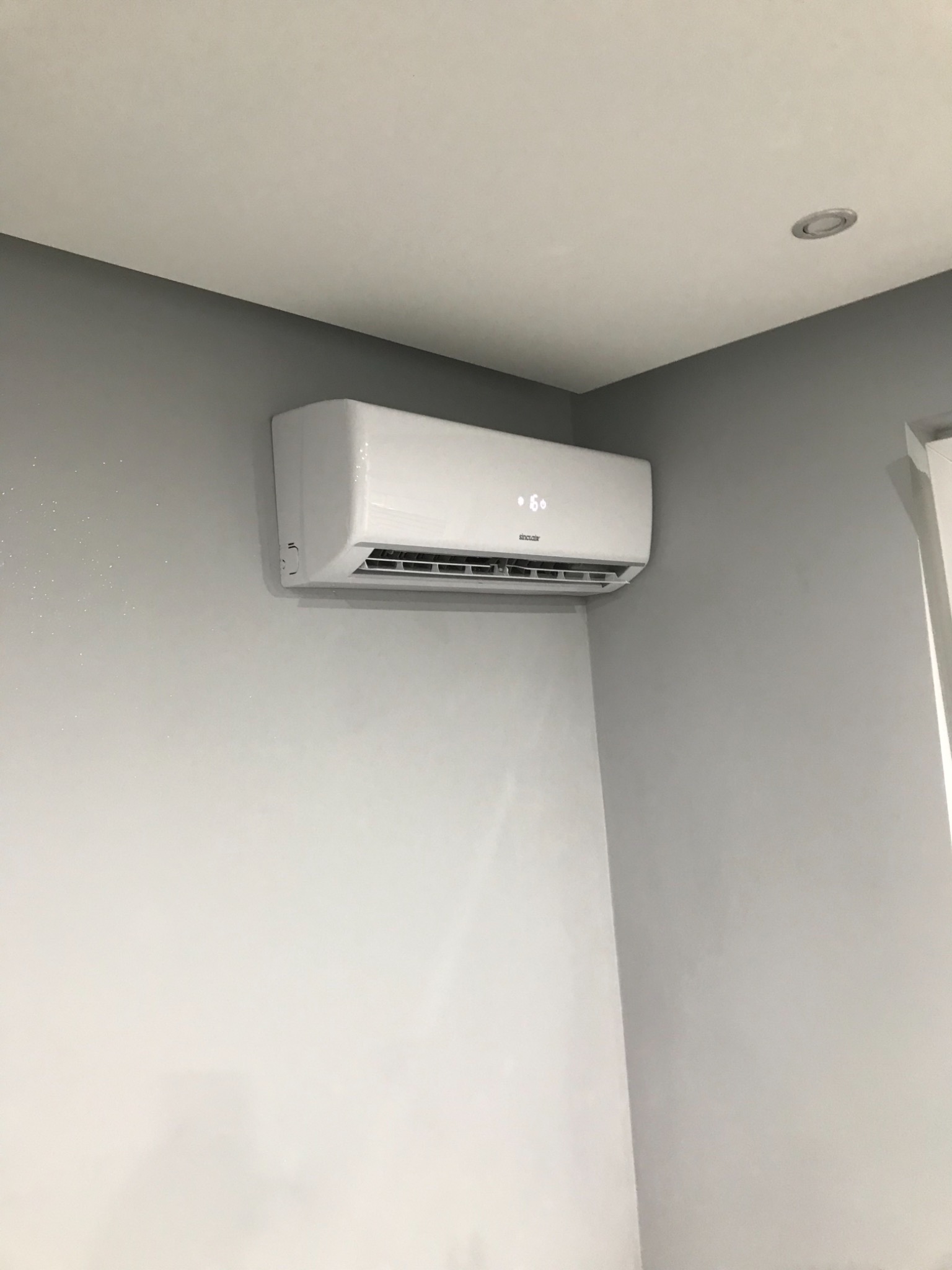 Biały klimatyzator marki Sinclair zamontowany na szarej ścianie w rogu pomieszczenia, widoczny wyświetlacz z temperaturą 16 stopni Celsjusza, w tle biały sufit z okrągłym oświetleniem.