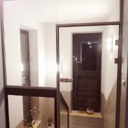 Szklana ściana oddzielająca klatkę schodową od wejścia, z przeszklonymi drzwiami na zawiasach z metalową ramą i klamką, widok na schody i rośliny doniczkowe.