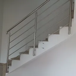 Schody z balustradą nierdzewną o minimalistycznym designie, widoczne mocowania do stopni, jasne ściany i podłoga z płytek w odcieniach beżu.