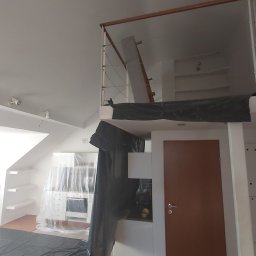 Yevhen Hurtovyi - Wnętrze mieszkania w trakcie remontu: kuchnia zabezpieczona folią malarską, widoczna antresola z drewnianą barierką i metalowymi linkami, brązowe drzwi wewnętrzne