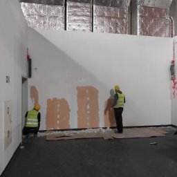 Yevhen Hurtovyi - Dwóch pracowników w żółtych kamizelkach i kaskach wyrównuje ścianę w pomieszczeniu z widocznymi instalacjami sufitowymi i zabezpieczeniami przeciwpożarowymi.