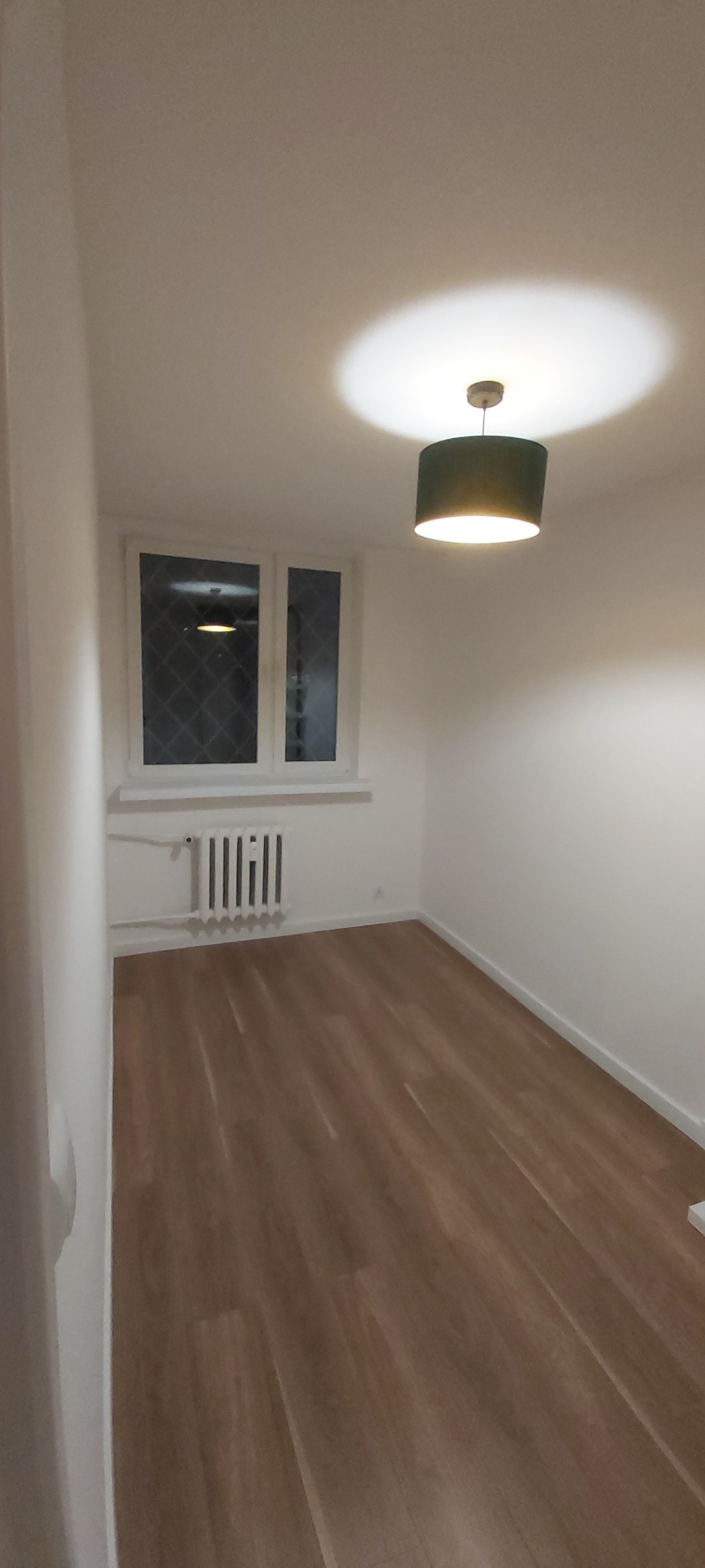 Pusty pokój po remoncie z panelami podłogowymi, białymi ścianami, grzejnikiem i oknem z kratką, oświetlony wiszącą lampą z zielonym abażurem.