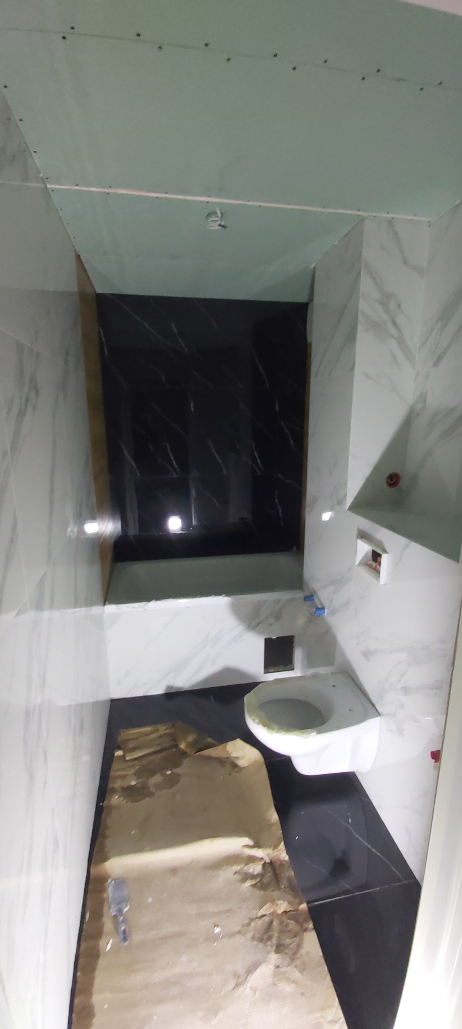 Wnętrze łazienki w trakcie remontu: białe marmurowe płytki ścienne, czarne płytki podłogowe, zamontowana toaleta, płyta karton gips na suficie, zabezpieczona podłoga.