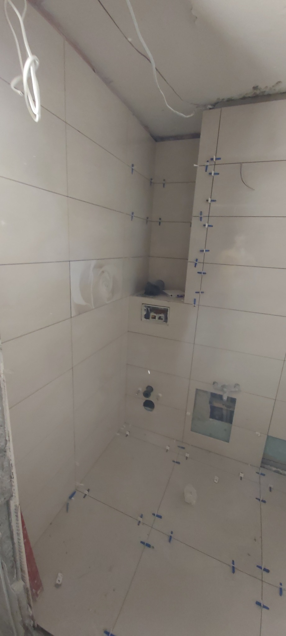 Ściany łazienki w trakcie układania jasnych, prostokątnych płytek z dekoracyjnym motywem kwiatowym. Widoczne krzyżyki dystansowe, przewody i otwory pod instalacje.