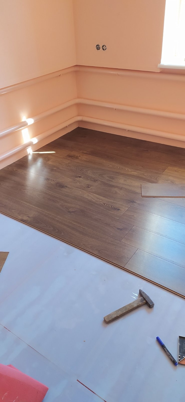 Układanie paneli podłogowych w narożniku pokoju, widoczne narzędzia: młotek i pisak, częściowo zabezpieczona podłoga folią ochronną.