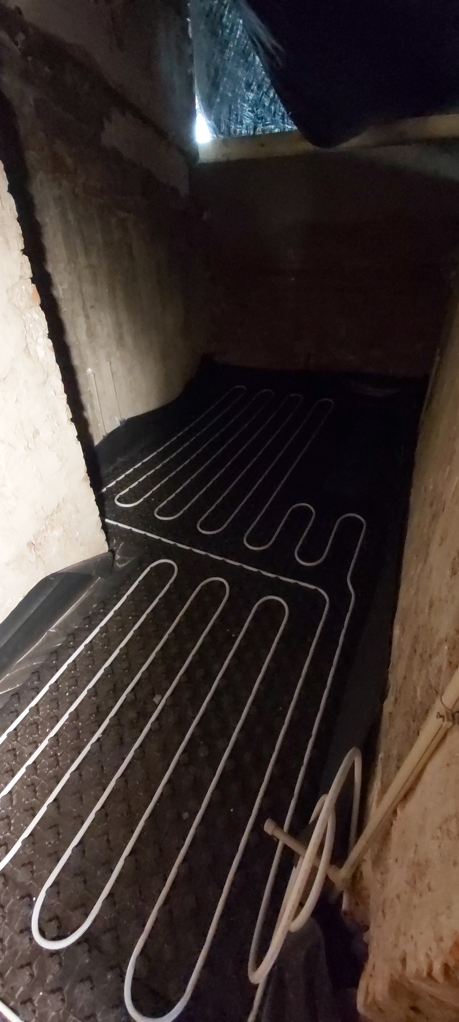 Widok z góry na system ogrzewania podłogowego z białymi rurkami ułożonymi na czarnej macie izolacyjnej, w surowym wnętrzu z betonowymi ścianami i widocznym fragmentem instalacji przy ścianie.