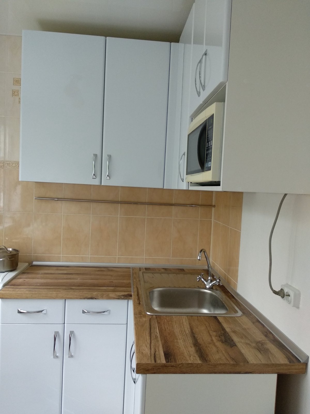 Jasna kuchnia z białymi szafkami, drewnianym blatem, beżowymi płytkami i metalowym zlewem. Na ścianie widoczny wieszak i gniazdko elektryczne.