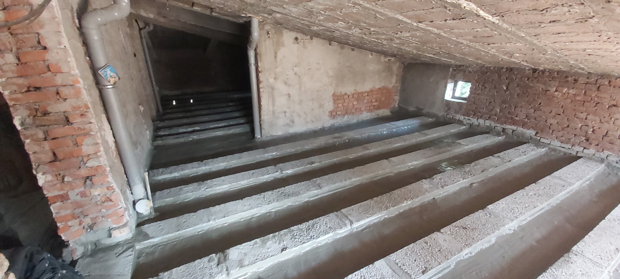 Surowy stan podłogi w remontowanym pomieszczeniu z ceglanymi ścianami, widoczne przygotowane podłoże pod wylewkę, pionowa rura kanalizacyjna przy ścianie.