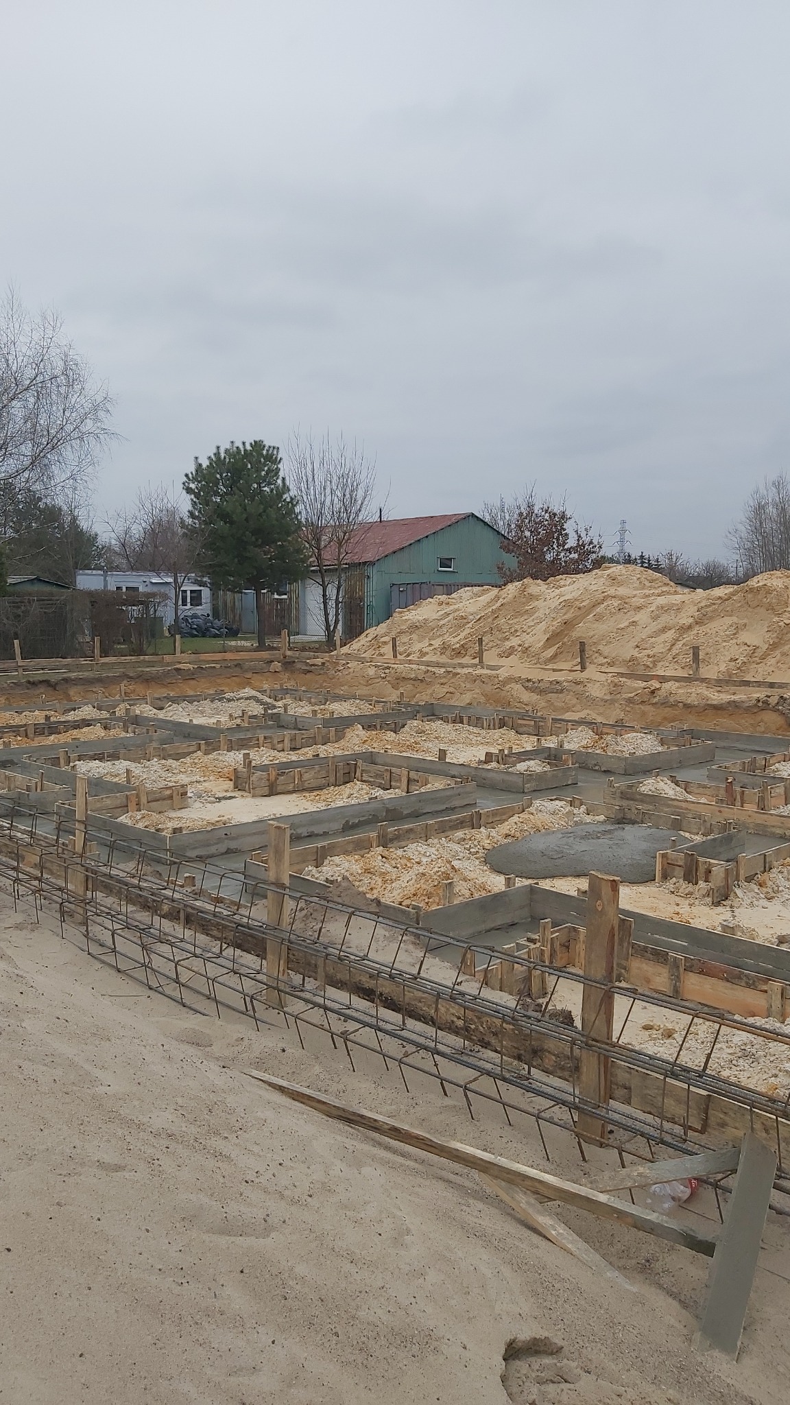 Wykop pod fundamenty z drewnianym szalunkiem i stalowym zbrojeniem, częściowo zalany betonem; w tle budynek gospodarczy i hałda piasku.