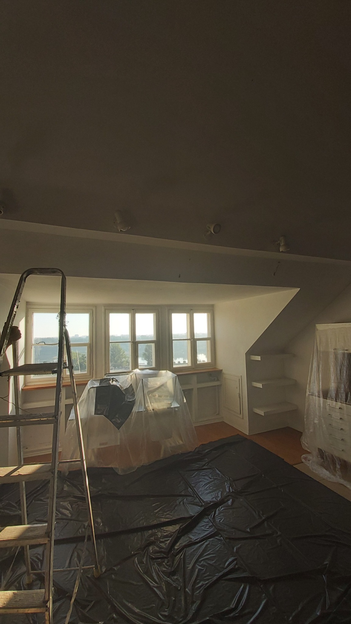 Pokój na poddaszu w Warszawie przygotowany do malowania: meble zabezpieczone folią, rozłożona folia ochronna na podłodze i drabina oparta o ścianę przy oknie.