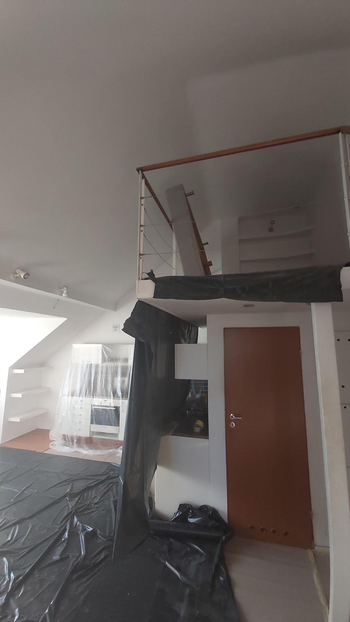 Wnętrze mieszkania w trakcie remontu: kuchnia zabezpieczona folią malarską, widoczna antresola z drewnianą barierką i metalowymi linkami, brązowe drzwi wewnętrzne