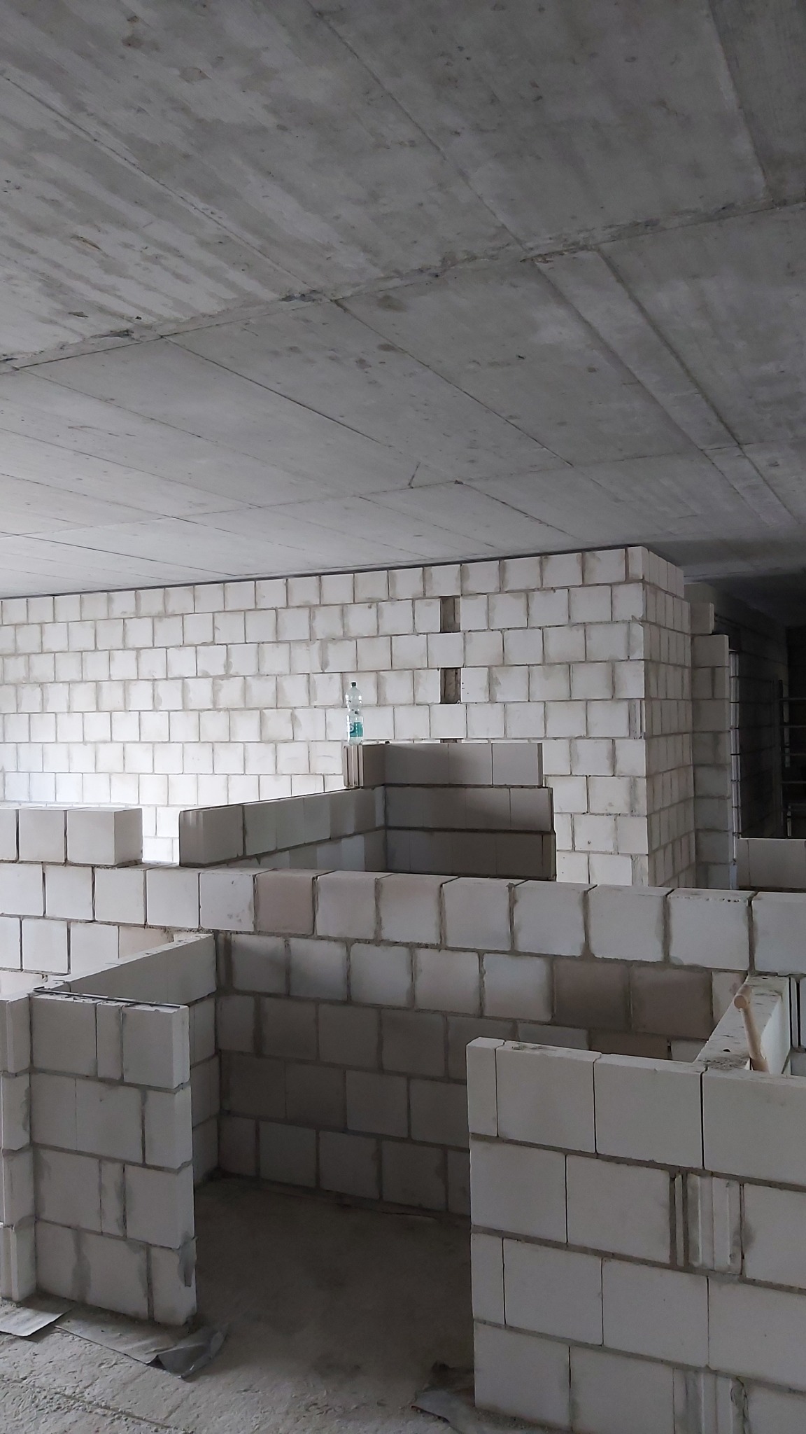 Wznoszone ściany działowe z białych bloczków, widoczna butelka wody na murze i surowy betonowy strop.