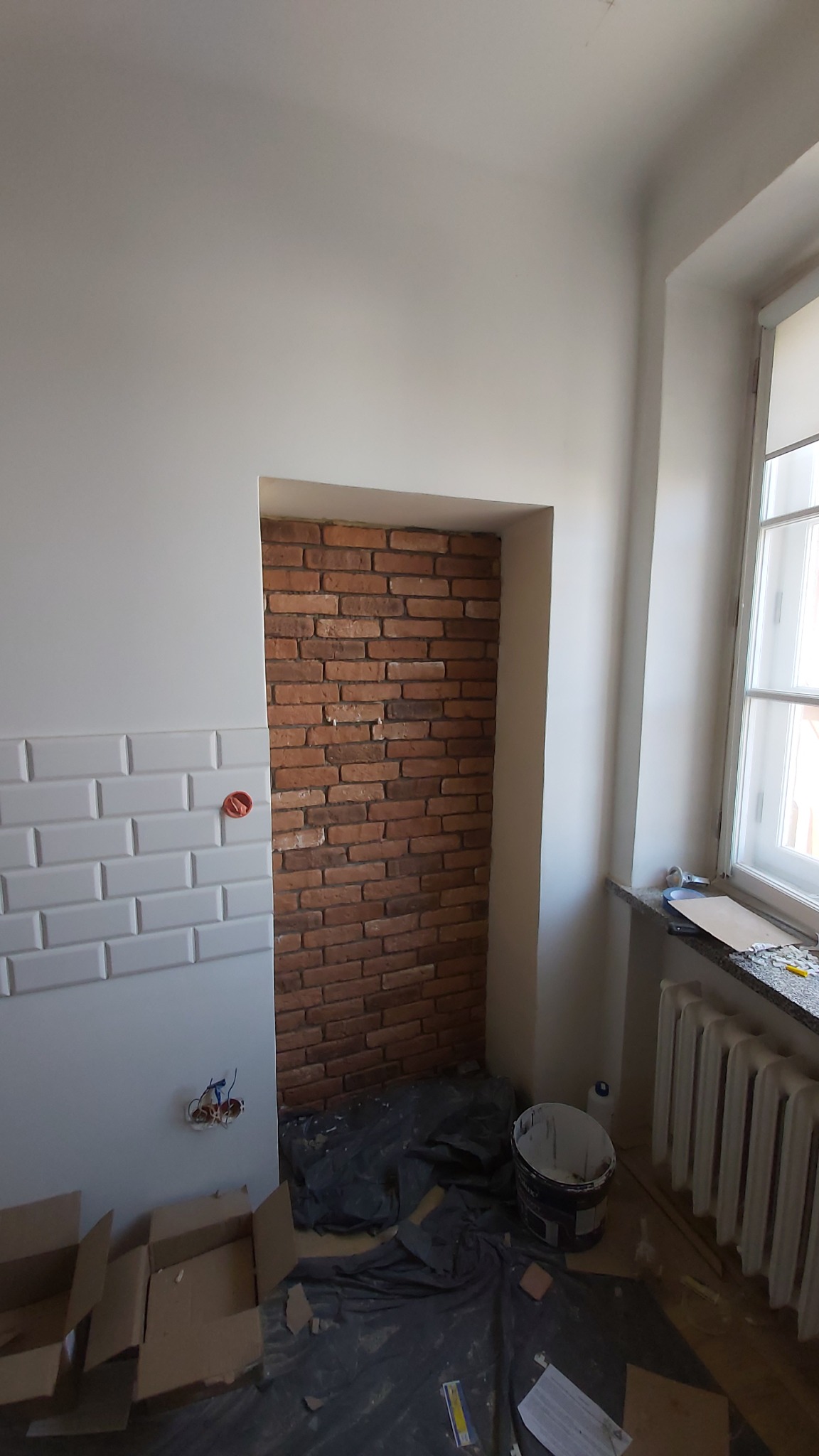 Częściowo wykończone wnętrze z ceglaną ścianą w niszy i białymi płytkami metra na sąsiedniej ścianie, podłoga zabezpieczona folią, widoczne puszki elektryczne i grzejnik.
