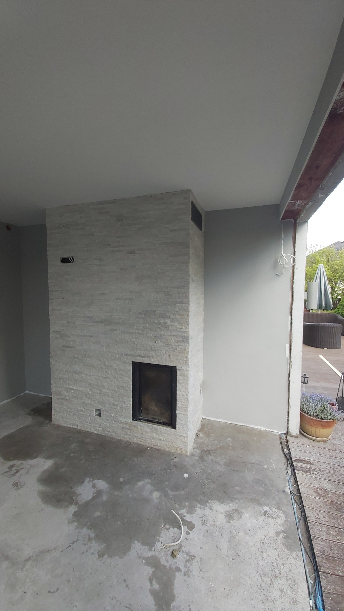 Wnętrze w trakcie remontu z kominkiem obłożonym jasną, nieregularną okładziną kamienną, surowy beton na podłodze i widoczne prace wykończeniowe.