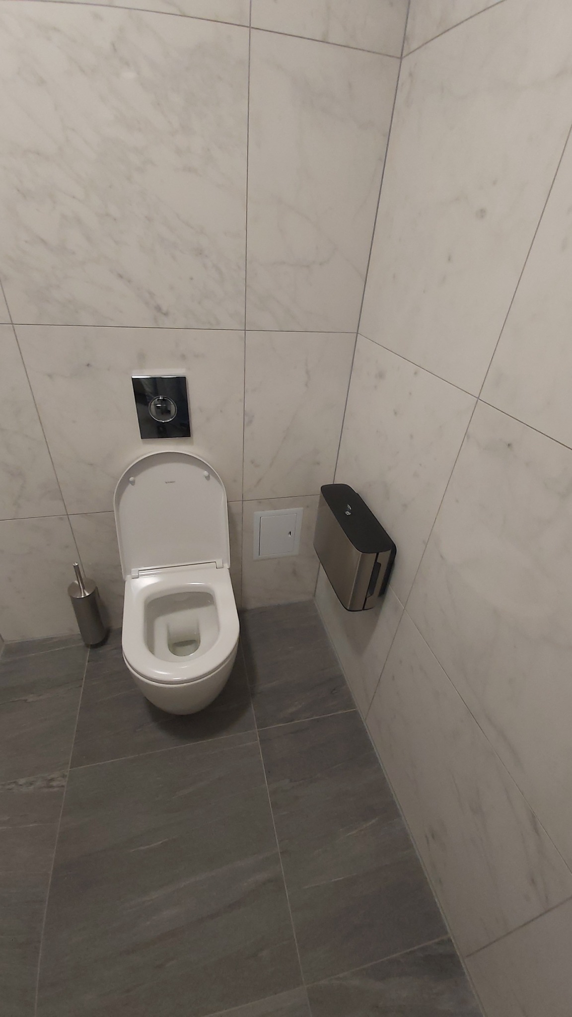 Nowoczesna łazienka z białą ceramiką, marmurowymi płytkami ściennymi i szarymi płytkami podłogowymi, widoczny szczotka do WC i metalowy pojemnik na papier toaletowy.