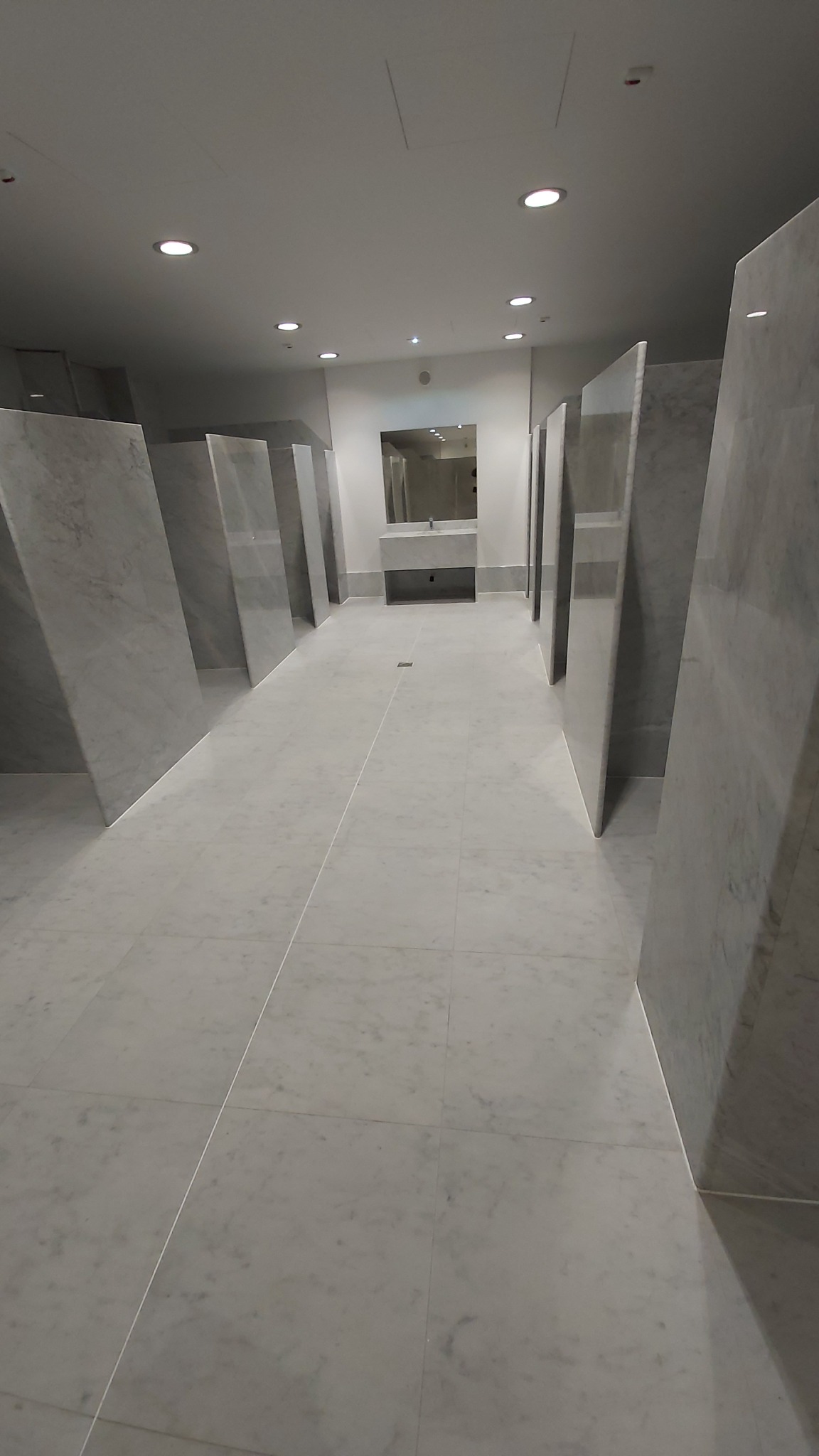 Wnętrze publicznej toalety wykończonej białym marmurem, z minimalistycznymi kabinami i umywalką w tle.