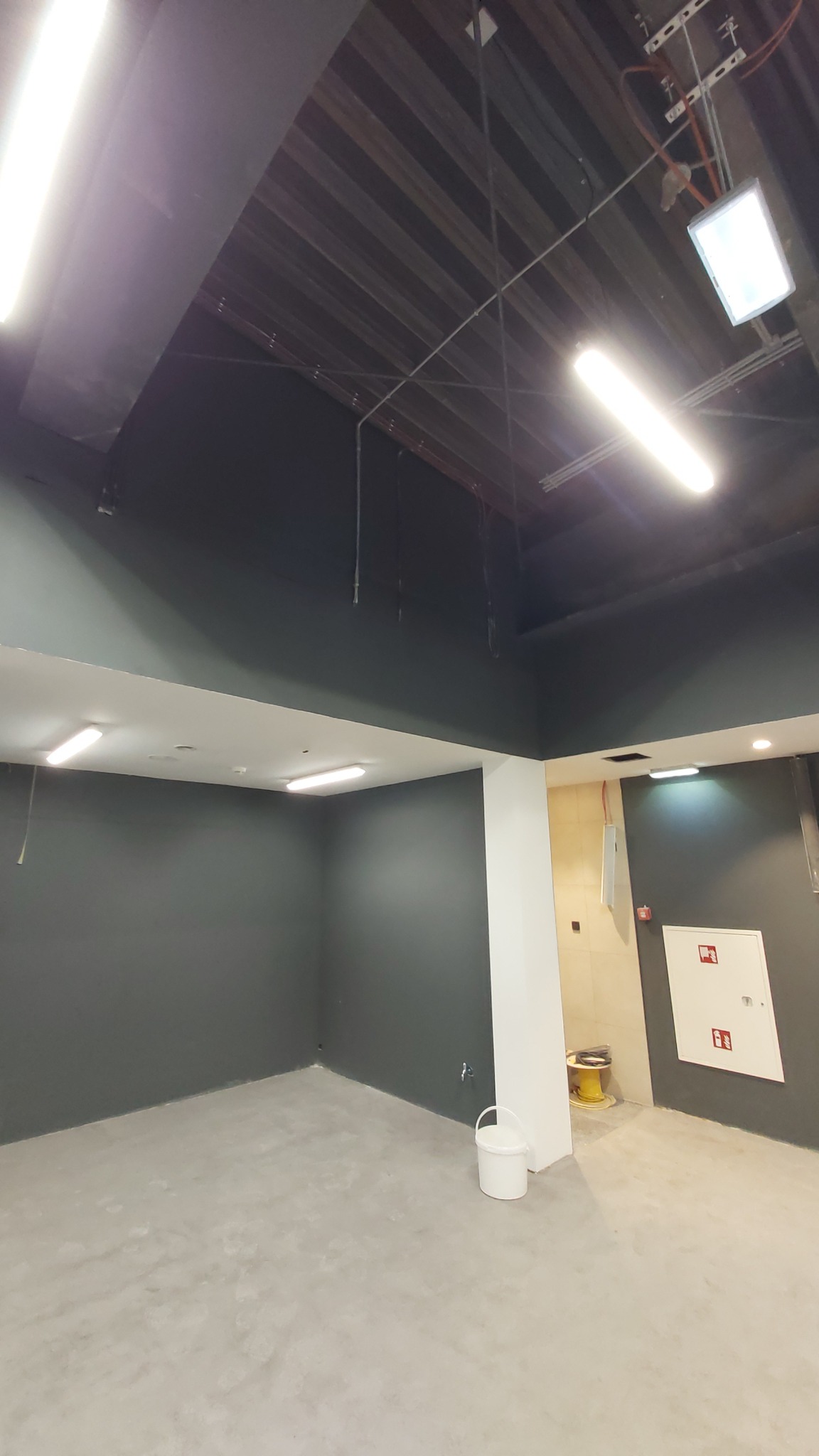 Surowe wnętrze biurowe z szarymi ścianami, betonową podłogą i odsłoniętym sufitem z instalacjami, widoczny biały pojemnik oraz żółta szpula z kablem w tle.